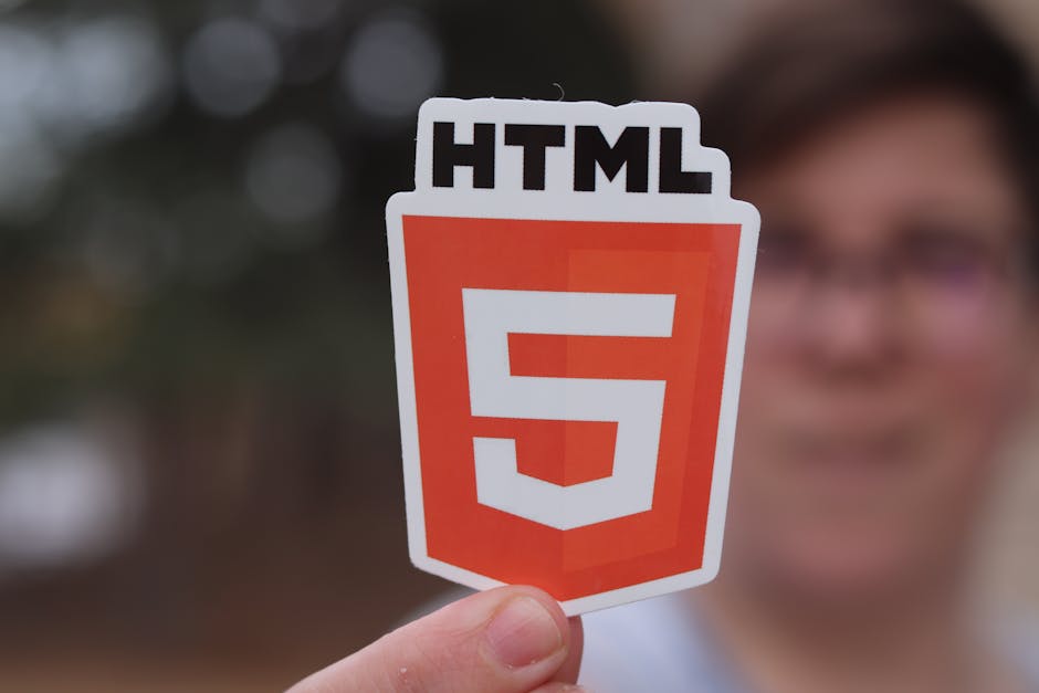 A person holding a laminated sign with the HTML5 logo prominently displayed in front of their face, which is slightly blurred, showing glasses and a smile. The background features an outdoor garden sc